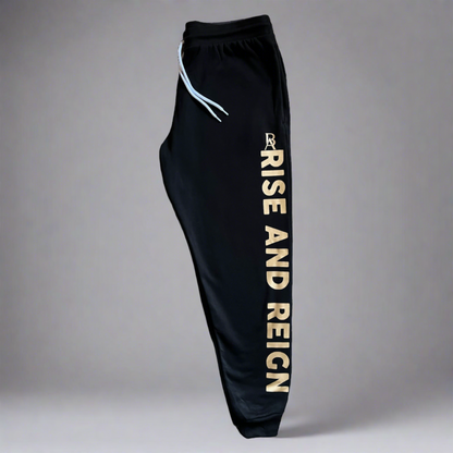Black 'Rise and Reign' joggers with gold print, featuring side pockets, ribbed ankle cuffs, and an adjustable white drawstring waistband. Made from a blend of Airlume combed and ring-spun cotton and polyester fleece.