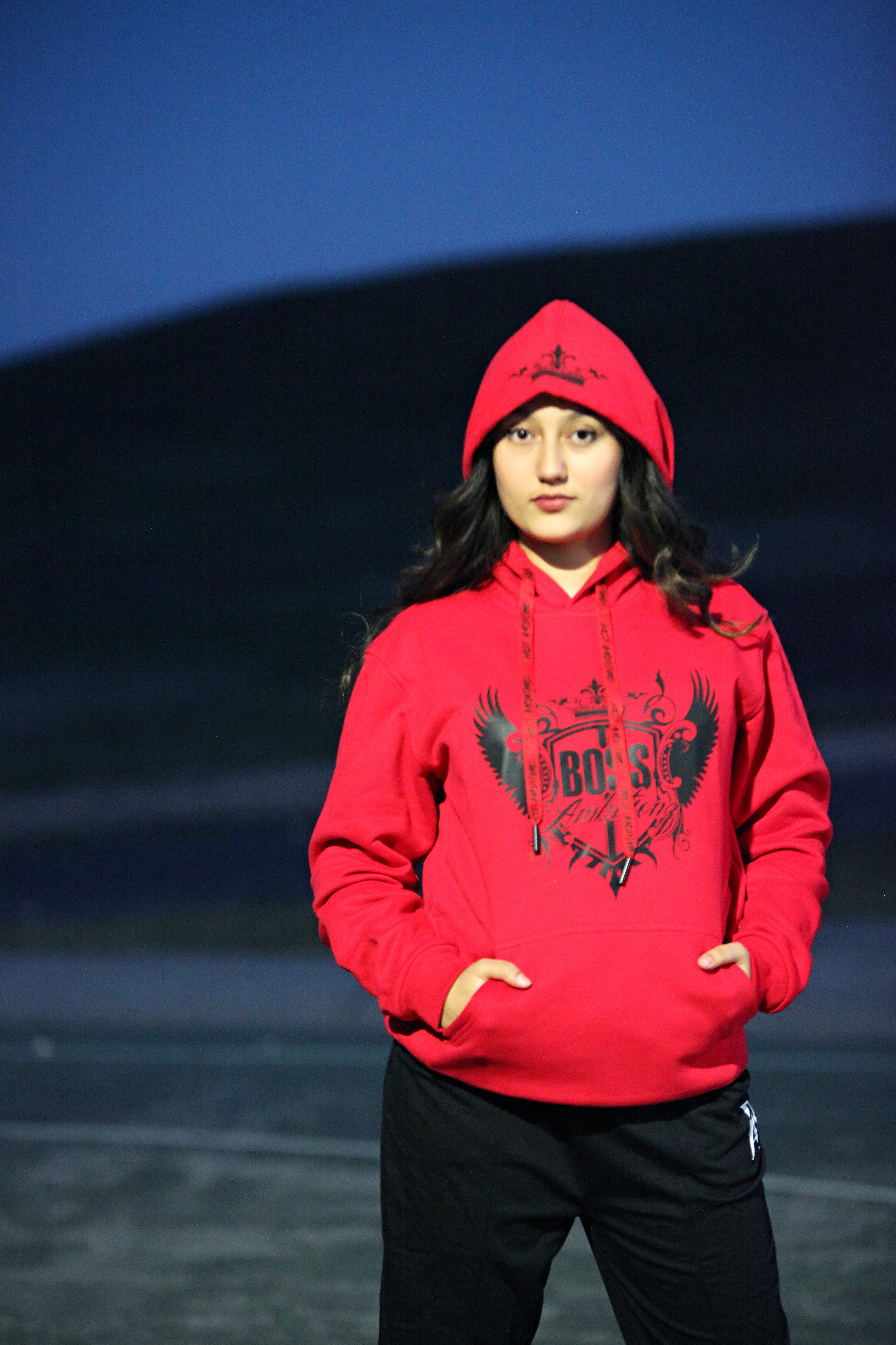 Miss Junior Teen USA models the Ambition Crown Radiance Reflective Hoodie in red, with the hood up, showcasing the reflective logo and confident stance.