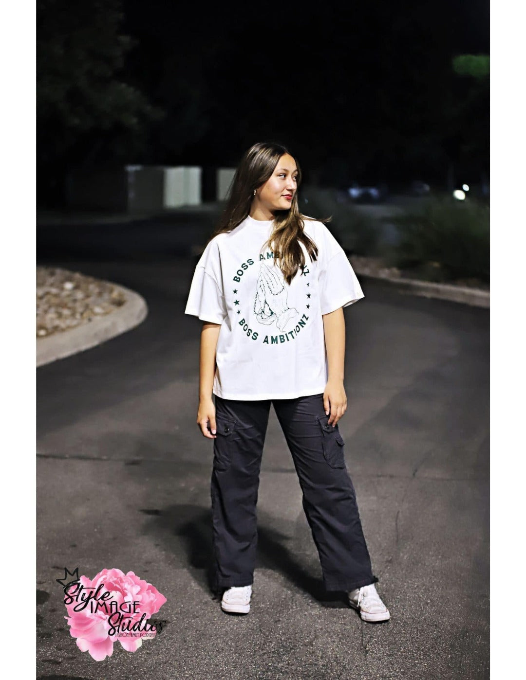 Full-body shot of Miss Junior Teen United USA in a relaxed pose on a dimly lit street, sporting a white Boss Ambitionz t-shirt with bold back print saying 'EVERYDAY ABOVE GROUND IS A GOOD DAY', paired with dark pants.