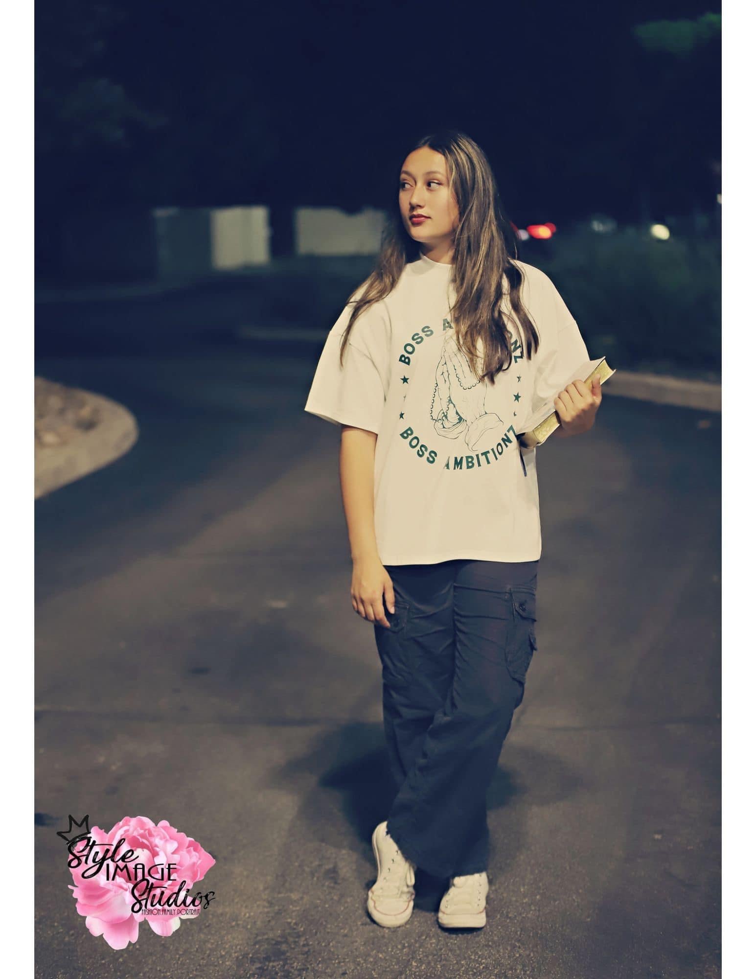 Miss Junior Teen United USA confidently models the Boss Ambitionz 'Everyday Luxe' oversized T-shirt on a night out. The front of the shirt features the distinctive 'Boss Ambitionz' hand graphic, while the back proclaims 'Everyday Above Ground is a Good Day' in bold letters. She pairs the tee with black cargo pants and white sneakers, embodying effortless street style