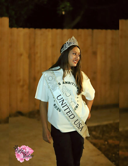 Miss Junior Teen United USA, celebrated pageant winner, showcases the Boss Ambitionz 'Everyday Luxe' Oversized T-Shirt. Her casual style is accentuated by the shirt's bold graphic, featuring an iconic prayer hands design, surrounded by the stars and script that read "BOSS AMBITIONZ". Perfect for those who blend laid-back fashion with a touch of luxury. Visit us at Boss Ambitionz Website to explore more. 