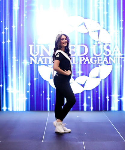 Rihana turning slightly on stage at the United USA National Pageant in Daytona Beach, FL, to showcase the side view of her outfit. She is wearing the Regal Lioness Tee from Boss Ambitionz, complemented by her sash and crown, embodying grace and ambition