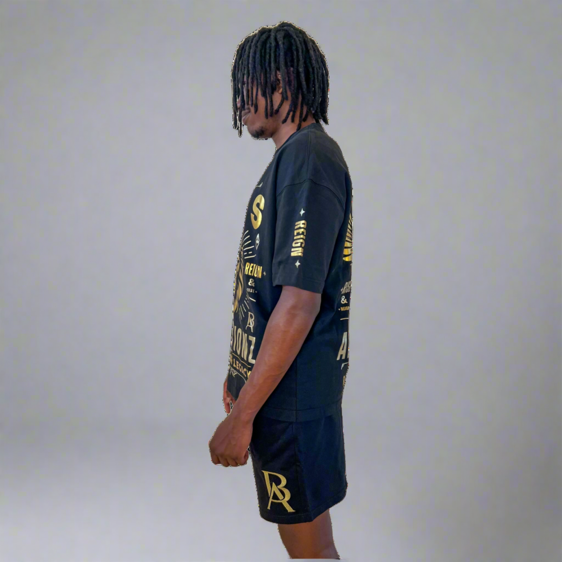 Side profile of DaKidswagg modeling Boss Ambitionz outfit: "Side profile of DaKidswagg modeling a black Boss Ambitionz t-shirt and shorts, showcasing the detailed gold text and logo designs