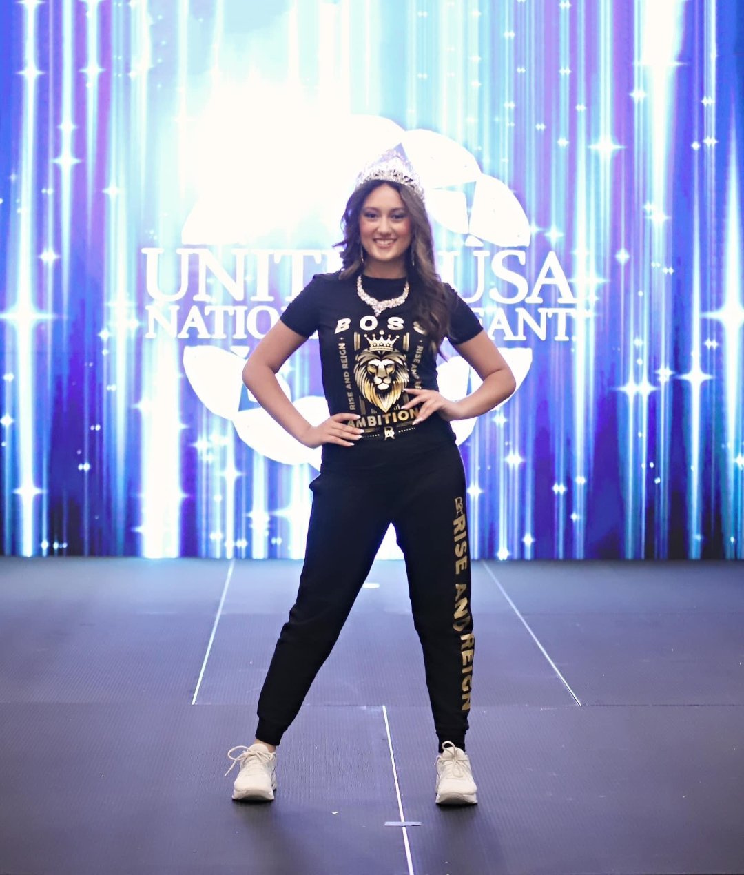 Rihana standing confidently on stage at the United USA National Pageant in Daytona Beach, FL, wearing the Regal Lioness Tee from Boss Ambitionz. She is smiling brightly, with her hands on her hips, showcasing the intricate lion design on her t-shirt."