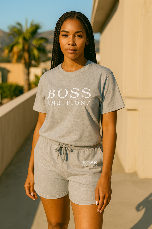 Woman wearing a gray 'BOSS AMBITIONZ' t-shirt and shorts on a balcony with palm trees in the background.