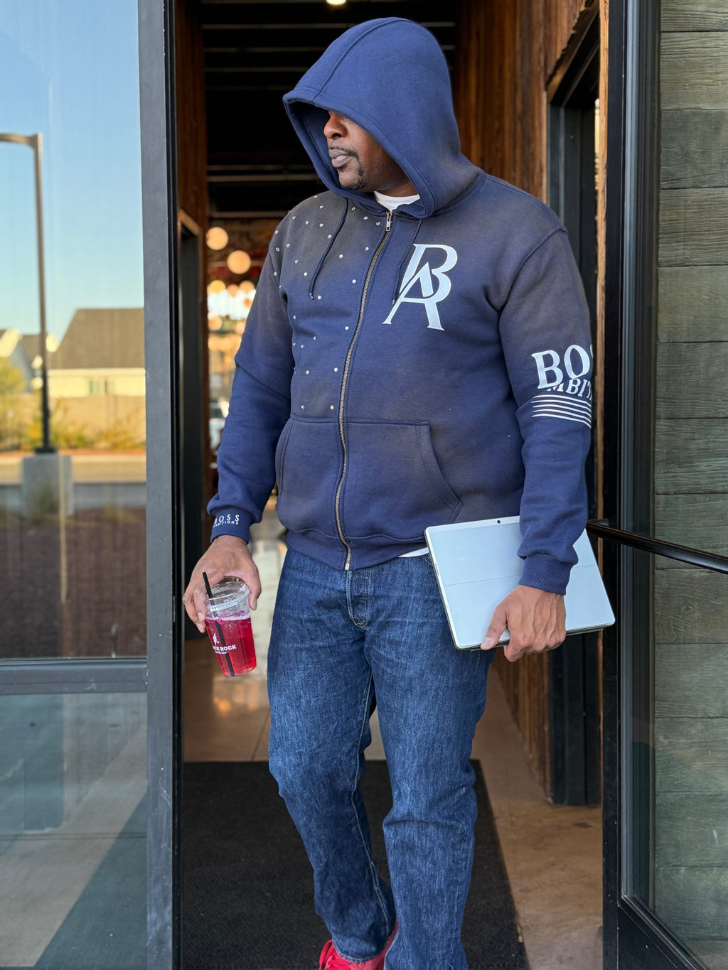 Man wearing a blue hoodie with a logo, holding a drink and a laptop, exiting a building.
