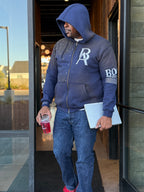 Man wearing a blue hoodie with a logo, holding a drink and a laptop, exiting a building.