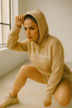 Woman wearing a tan 'BOSS' hoodie and shorts in a minimalistic setting.