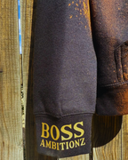 Close-up of the Rustic Hustlers Rhinestone Hoodie sleeve showing gold “BOSS AMBITIONZ” print on a distressed black-and-brown acid wash fabric with sunlight highlighting the texture.