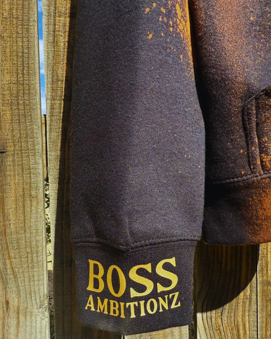 Close-up of the Rustic Hustlers Rhinestone Hoodie sleeve showing gold “BOSS AMBITIONZ” print on a distressed black-and-brown acid wash fabric with sunlight highlighting the texture.