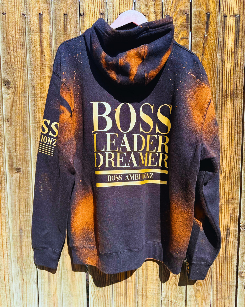Back view of the Rustic Hustlers Rhinestone Hoodie showing bold gold lettering that reads “BOSS LEADER DREAMER” and “BOSS AMBITIONZ,” on a black hoodie with orange acid-wash pattern and distressed finish.