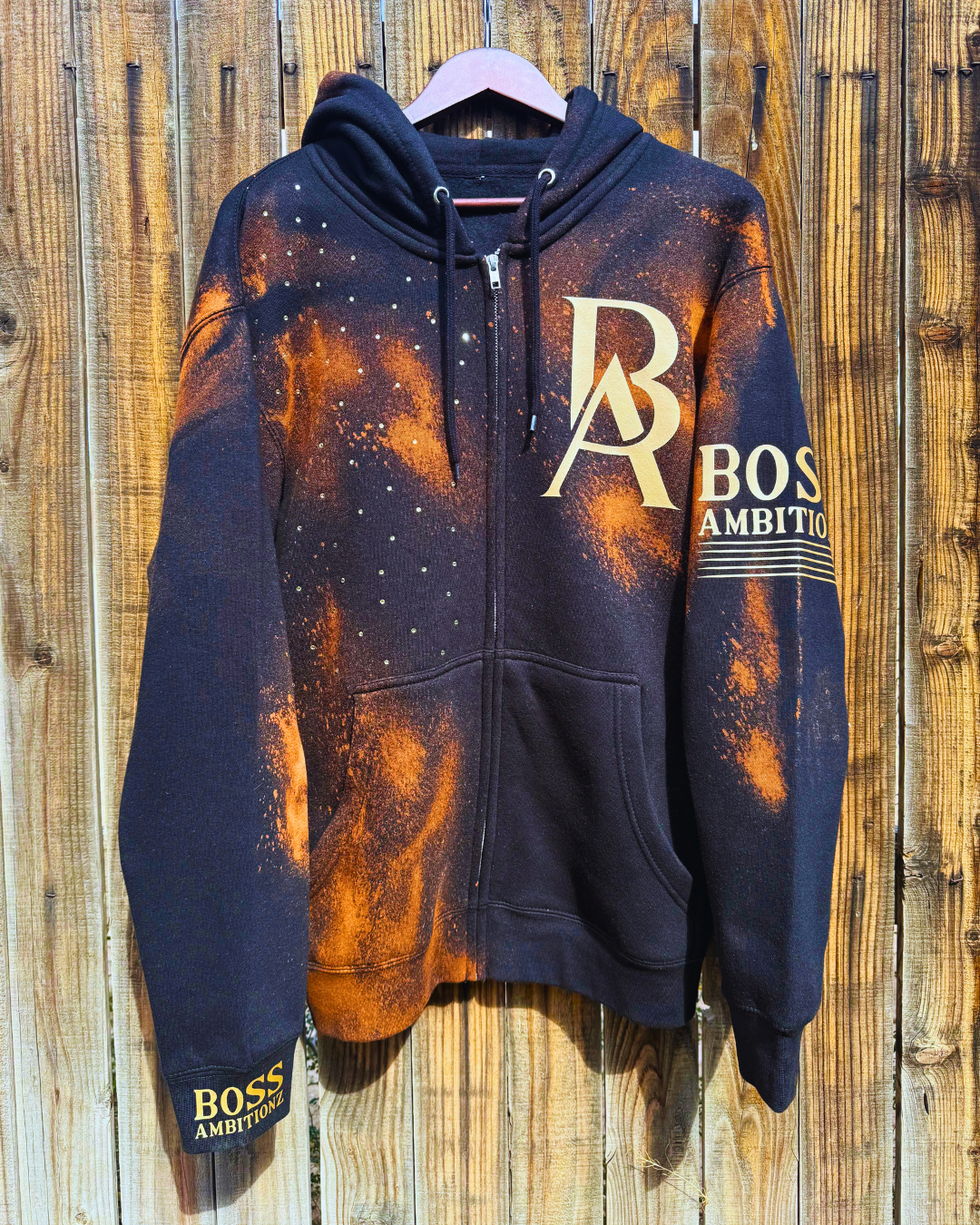 Front view of the Rustic Hustlers Rhinestone Zip-Up Hoodie by Boss Ambitionz, featuring an orange acid-wash finish on black fleece with gold BA logo, rhinestone detailing on the chest, and sleeve branding that reads “BOSS AMBITIONZ.
