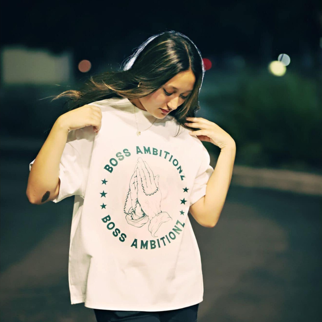 Miss Junior Teen United USA with a playful expression, wearing a white oversized Boss Ambitionz t-shirt with a graphic of praying hands encircled by stars and text, styled casually with dark cargo pants